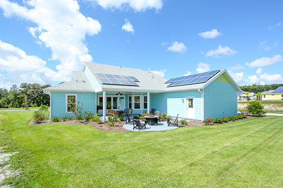 Single-story light blue home with solar panels and a patio, surrounded by green lawn.