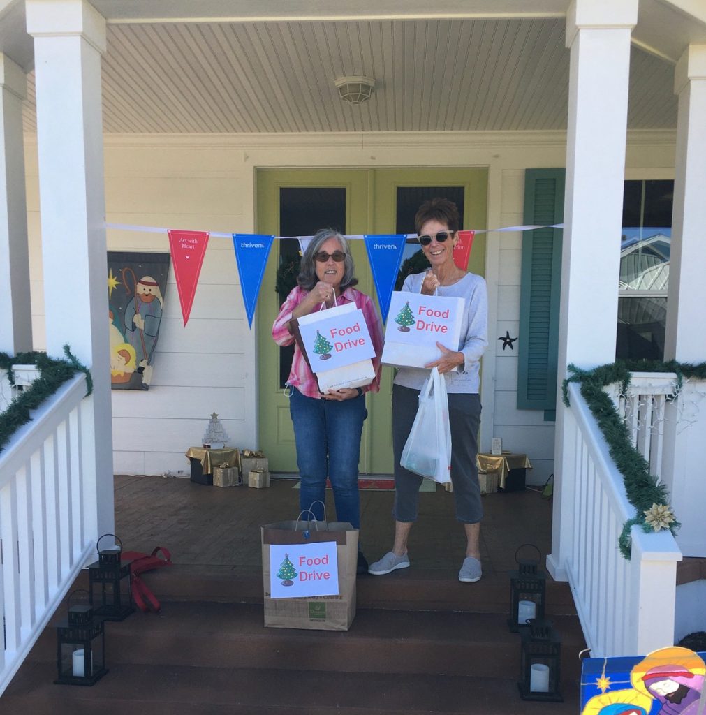 Two women on a front porch holding Food Drive donation bags, with holiday decorations and a nativity scene nearby.