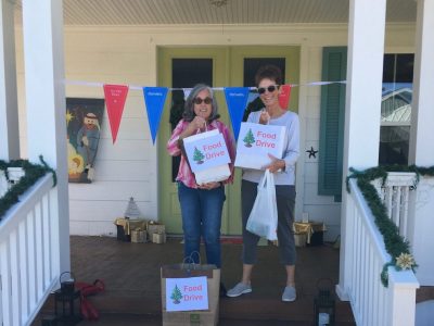 Two women on a front porch holding Food Drive donation bags, with holiday decorations and a nativity scene nearby.
