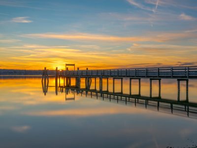 Sunset over calm water with a wooden pier reflecting golden light in the sky.