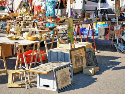 Outdoor flea market tables display vintage brass clocks, framed art, and collectibles on a sunny day.