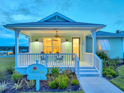 Front porch of the Sand Dollar model home with white railings, green siding, and a lit interior at dusk.