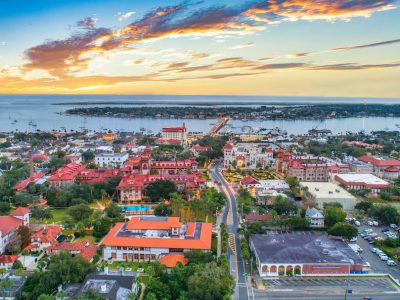 Aerial view of St. Augustine at sunset, featuring red-roofed buildings, a bridge, and boats on the water.