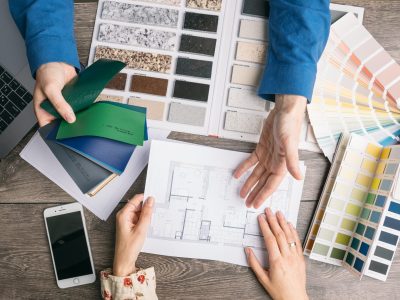 Overhead view of hands reviewing home floor plans, color swatches, and material samples on a wooden desk.