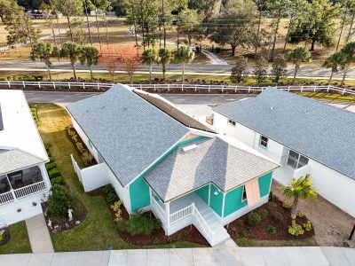 Aerial view of a teal and white home with a gray shingled roof and solar panels, surrounded by green landscaping.