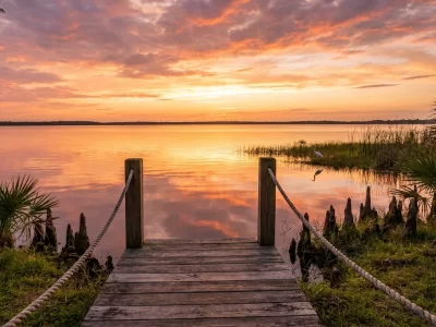 Late golden hour over a still Central Florida lake, seen from a weathered wooden dock with cypress knees and saw palmettos along the bank under a wide warm sky